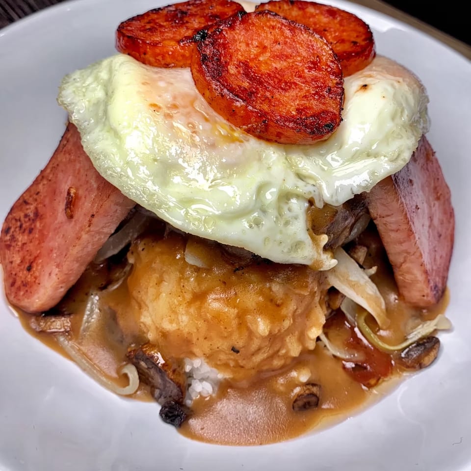 A close-up of a Hawaiian loco moco dish with rice, gravy, Spam, sausage, and a fried egg.